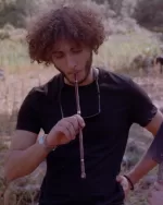 Young man with curly hair in a black T-shirt outdoors, holding a long smoking pipe to his mouth with sunglasses hanging from his neck.