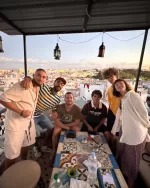 Six friends on a rooftop terrace with a cityscape backdrop at sunset or golden hour.