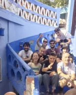 Group of smiling friends posing for a photo on a bright blue-painted outdoor staircase, some waving at the camera.